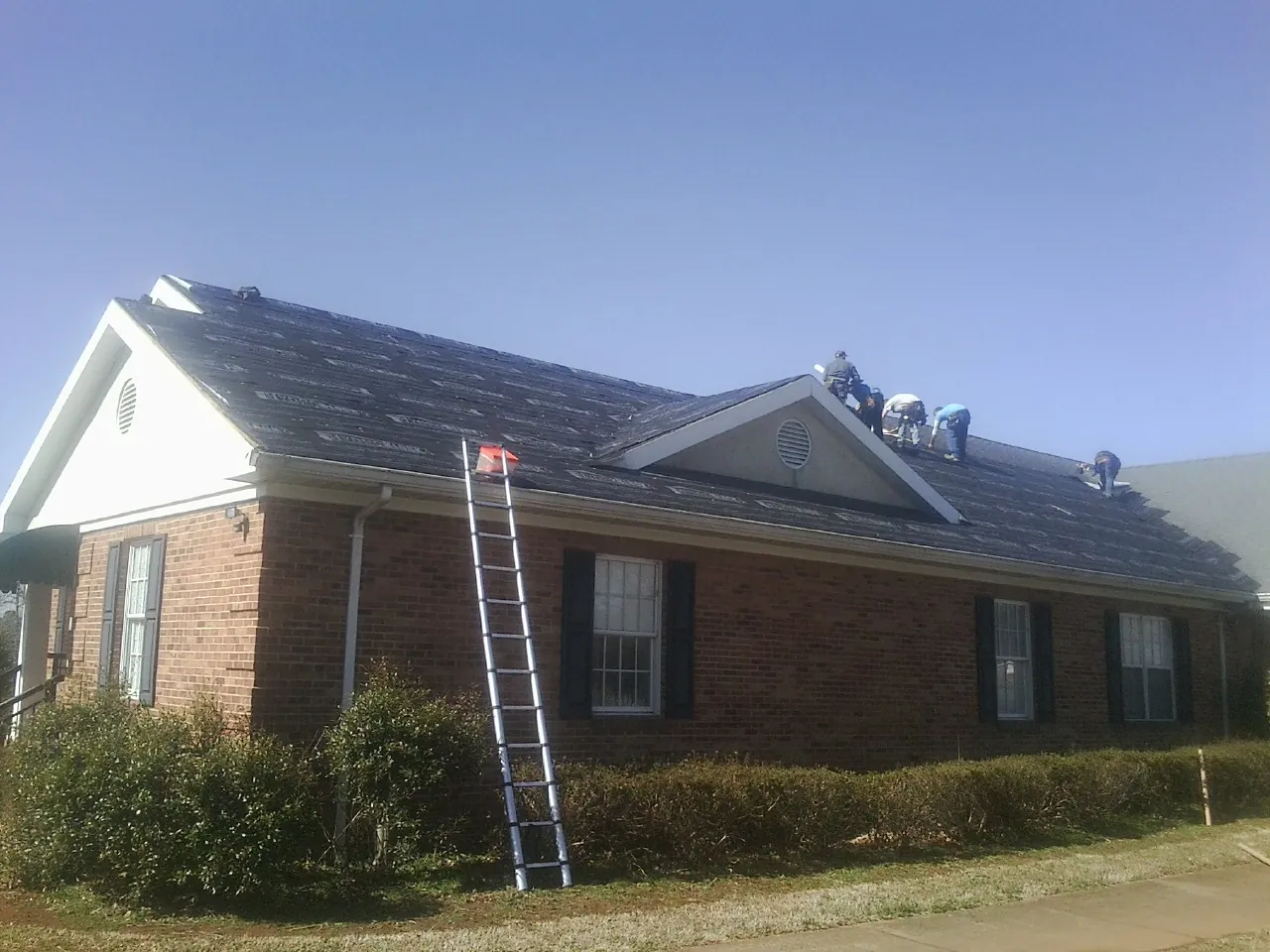 Crew tearing shingles off a brick ranch roof with a ladder set — Metal Roof Repair work by our crew in Sand Lake